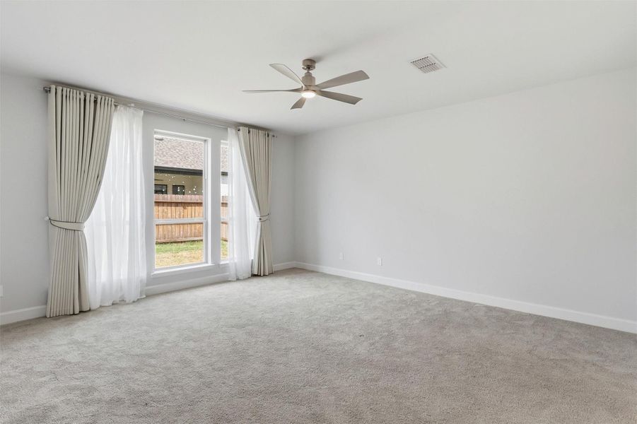 The expansive primary suite offers a peaceful retreat with plush carpeting, a designer ceiling fan, oversized windows dressed with elegant custom drapery, and plenty of space for a complete bedroom suite and sitting area.