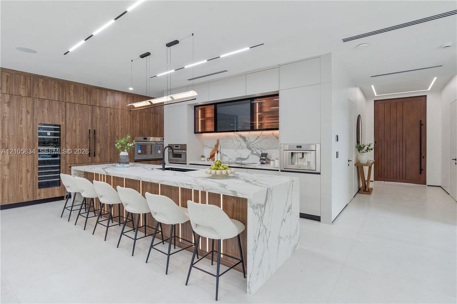 Modern kitchen featuring flat-panel wood-finish cabinetry, a large waterfall island with stone countertop, built-in stainless steel appliances, integrated under-cabinet lighting, and recessed linear ceiling illumination