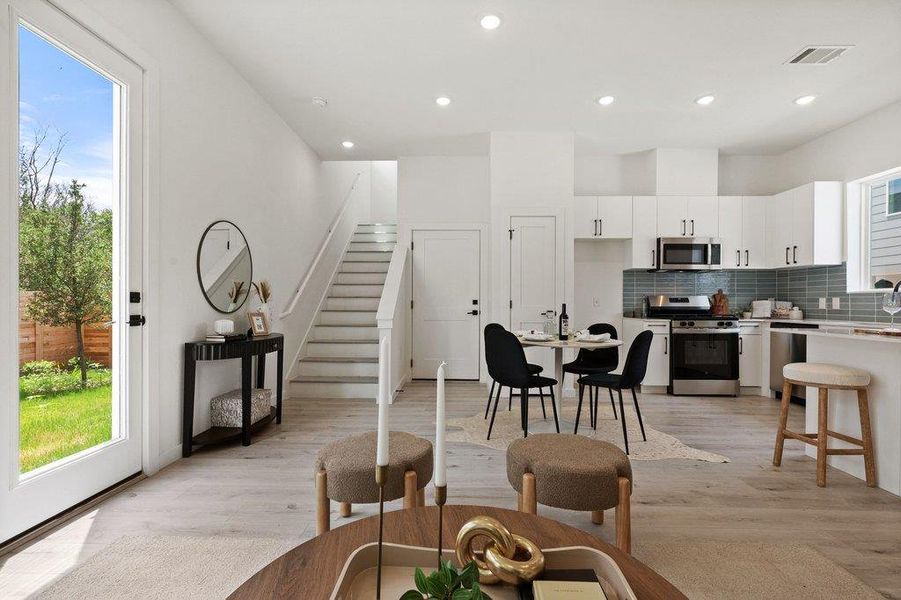 Open-concept living area featuring light-colored flooring, a kitchen with white cabinetry and stainless steel appliances, and a staircase with white risers and handrails