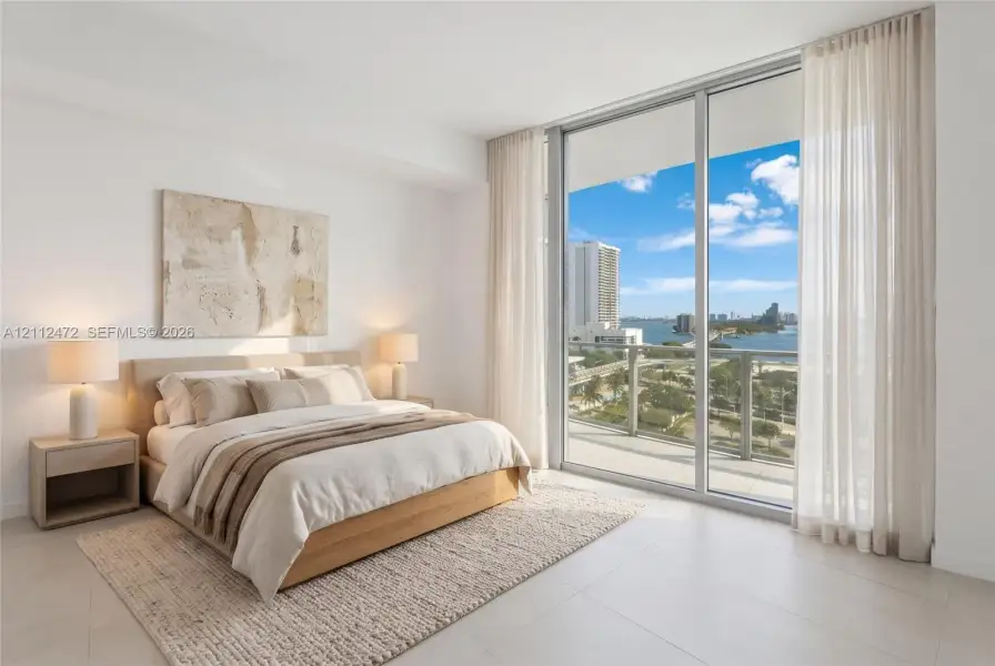 Floor-to-ceiling glass sliding doors opening to a private balcony with water and skyline surroundings, neutral large-format tile flooring, and crisp white wall finishes. VIRTUALLY STAGED