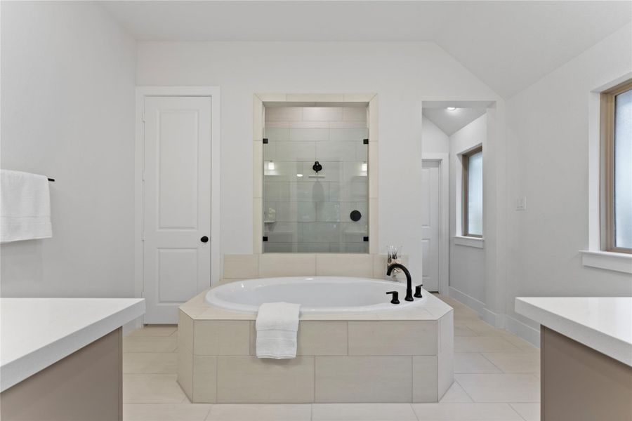 Spacious bathroom featuring a built-in soaking tub with black fixtures, a walk-in glass-enclosed shower, dual vanity countertops, and light-toned tile flooring