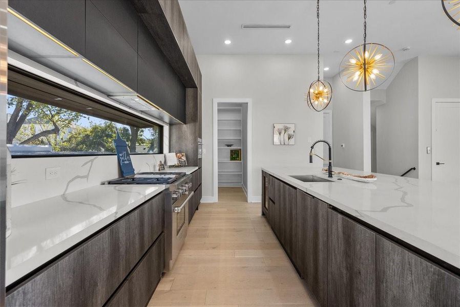 This modern kitchen features sleek dark cabinetry, light wood floors, and an expansive white countertop with a built-in sink. Large windows provide ample natural light, while contemporary pendant lights add elegance. A spacious pantry is accessible through a nearby door. This modern kitchen features sleek dark cabinetry, light wood floors, and an expansive white countertop with a built-in sink. Large windows provide ample natural light, while contemporary pendant lights add elegance. A spacious pantry is accessible through a nearby door.