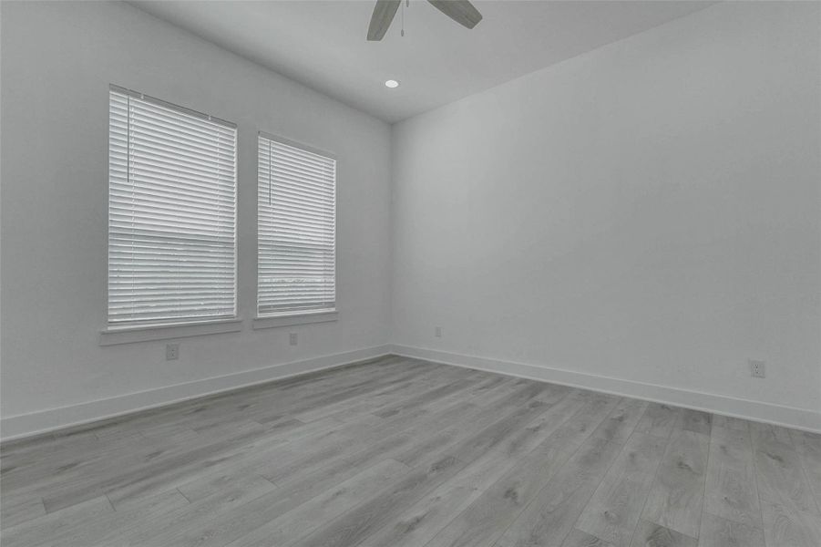 The upstairs primary bedroom is filled with natural light from two tall windows and finished with light wood-look flooring, recessed lighting, a ceiling fan, high ceilings, and white blinds. The spacious neutral setting creates a calm private retreat.