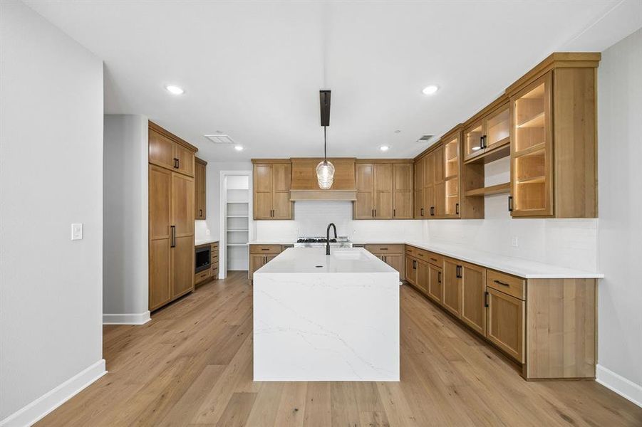 Kitchen with a center island with sink, light stone counters, hanging light fixtures, wood finish cabinetry, and decorative backsplash Kitchen with a center island with sink, light stone counters, hanging light fixtures, wood finish cabinetry, and decorative backsplash