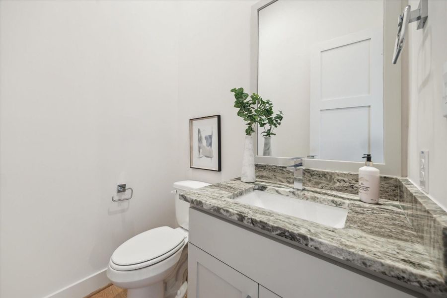 This photo shows a modern powder room featuring a sleek marble countertop, an undermount sink, and contemporary fixtures. The space is bright and well-lit, with a large mirror and minimalistic decor, including a small piece of framed art and a plant for a touch of greenery.