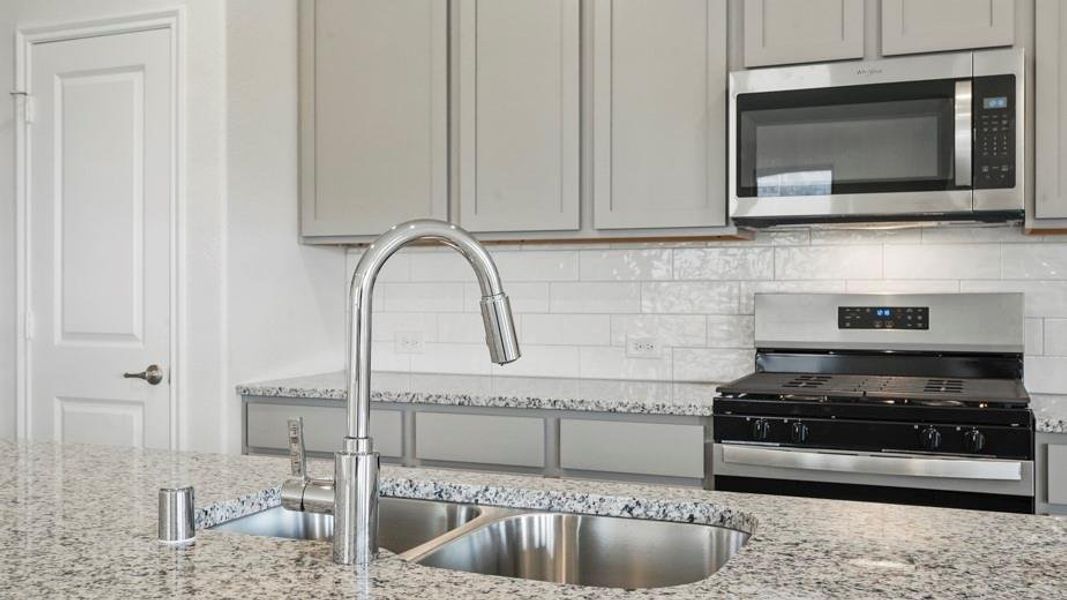 Kitchen featuring a double basin stainless steel sink with a high-arch faucet, granite countertops, gray cabinetry, white subway tile backsplash, and stainless steel appliances including a microwave and range
