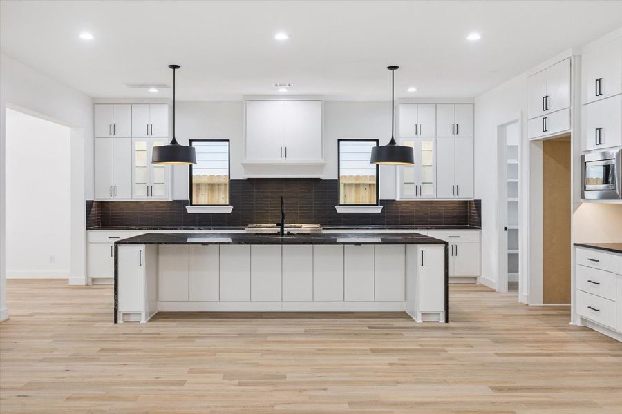 This designer kitchen makes a bold statement with its waterfall-edge island, black countertops, and contrasting pendant lights. Abundant cabinetry, dual windows, and a walk-in pantry offer both beauty and function, perfect for cooking, gathering, and entertaining. This designer kitchen makes a bold statement with its waterfall-edge island, black countertops, and contrasting pendant lights. Abundant cabinetry, dual windows, and a walk-in pantry offer both beauty and function, perfect for cooking, gathering, and entertaining.