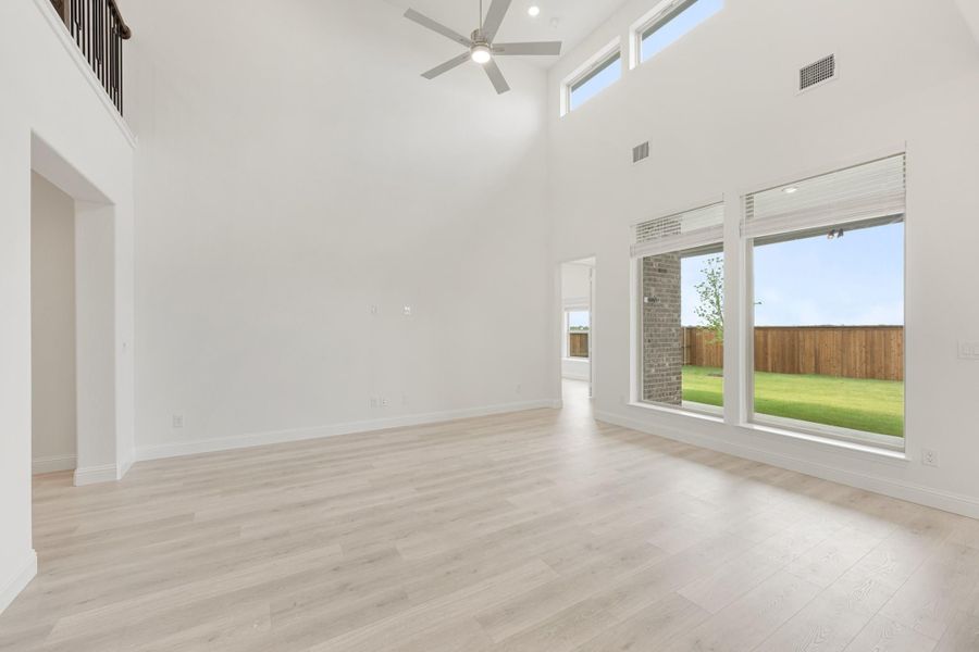 Open living room with two-story ceiling, ceiling fan, light wood floors, and large windows overlooking backyard