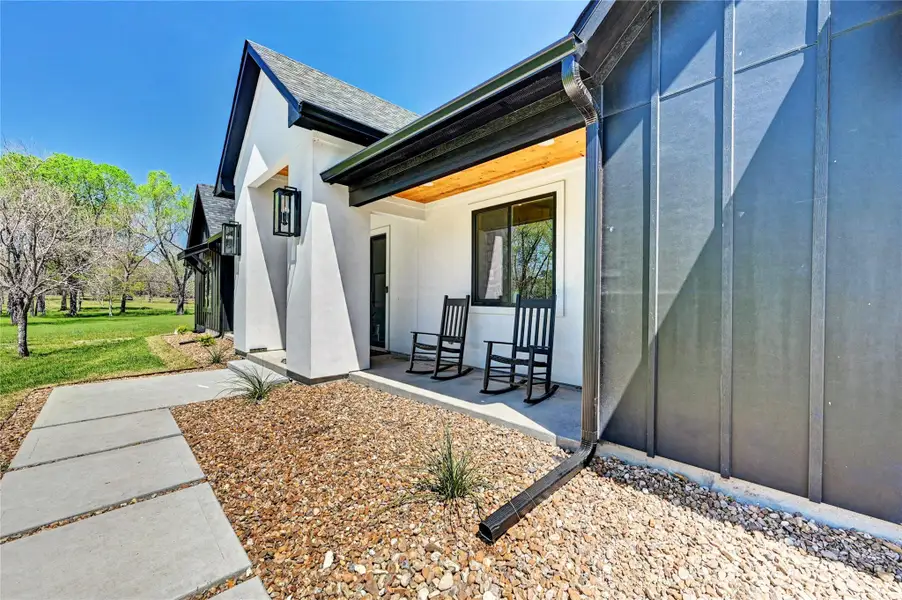 This photo showcases a modern, inviting home exterior with a combination of white and dark siding. The porch features two rocking chairs and overlooks a landscaped area with stone accents, surrounded by lush greenery.