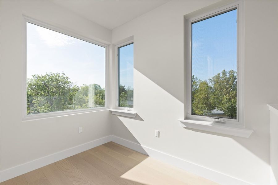 Sunlight pours across pale hardwood floors through large, clear windows overlooking mature trees.