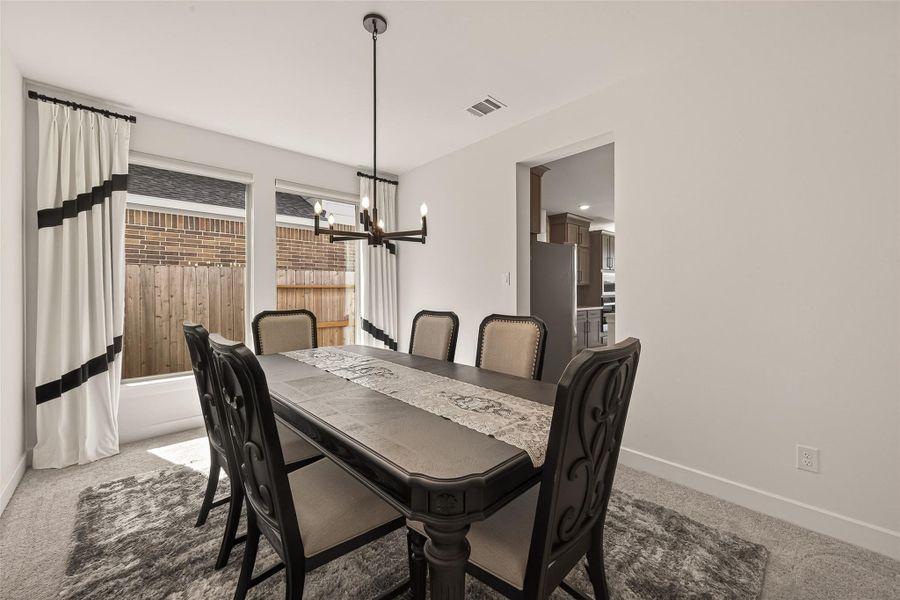 This is a gorgeous dining room with a modern chandelier, a dark wood table, and elegant chairs. Large windows offer natural light, while the neutral walls and carpet create a warm atmosphere. A doorway leads to a kitchen with stainless steel appliances.