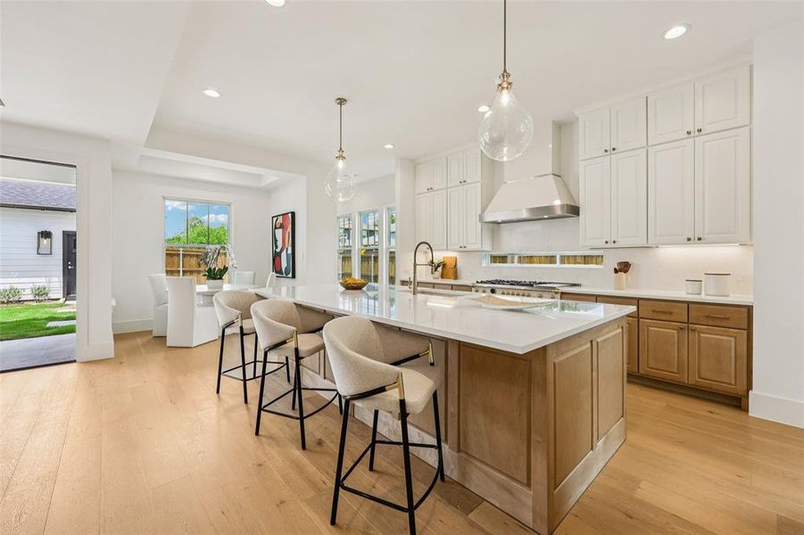 Contemporary kitchen featuring a spacious island with a light-toned countertop, a stainless steel range hood, white upper cabinetry, and natural wood-tone lower cabinetry