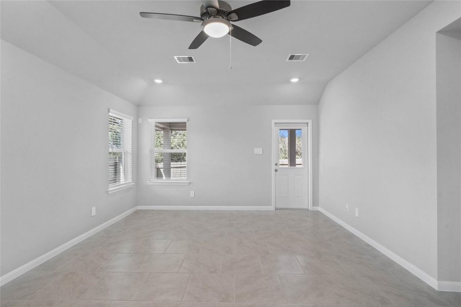 A bright and airy open-concept space featuring tile flooring, sloped ceilings with recessed lighting and ceiling fan, abundant natural light from large windows (fitted with blinds), and an access door to the back covered patio.