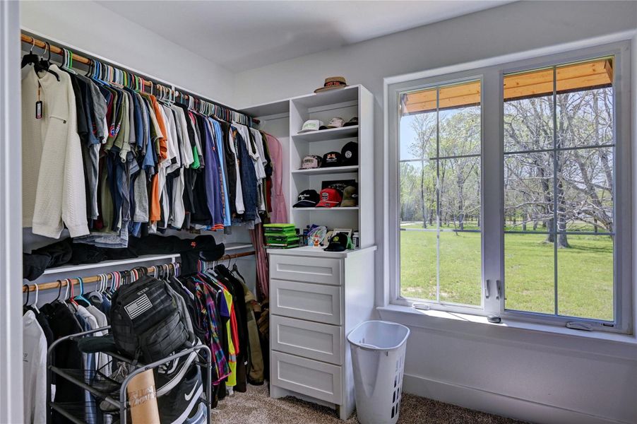 This photo showcases a spacious walk-in closet with ample hanging space, shelving, and drawers. Large windows provide natural light and offer a serene view of a green yard. Perfect for organizing and enjoying a bright, airy atmosphere.