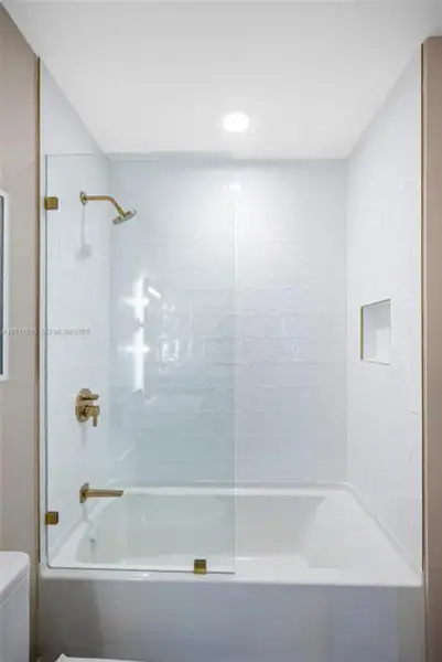Bathtub and shower combination featuring white textured wall tile, glass shower panel with gold-tone hardware, recessed shower niche, and gold-tone wall-mounted faucet and shower head