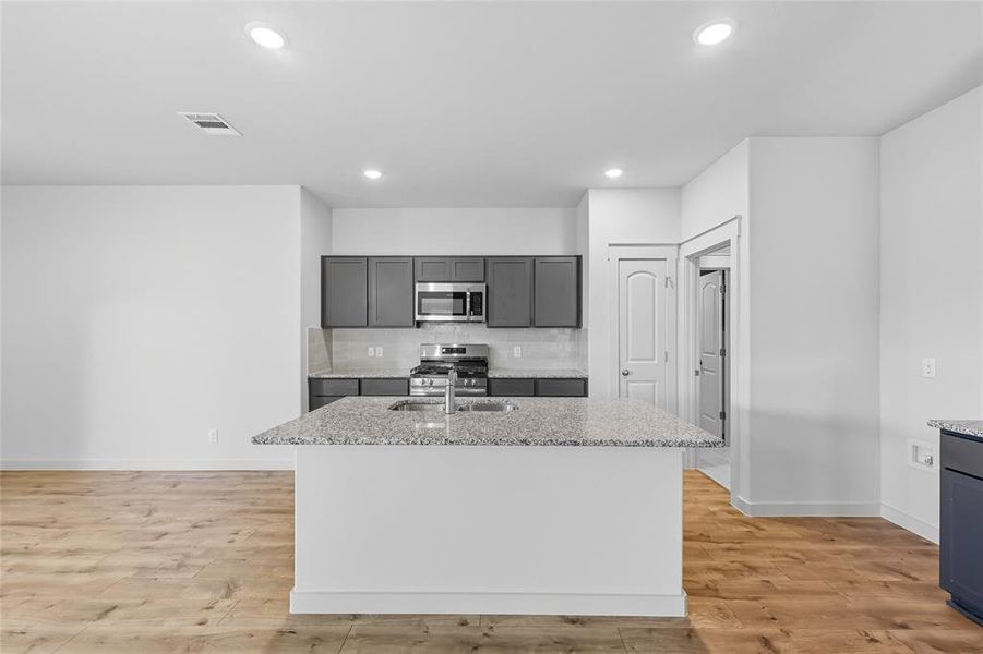 Kitchen featuring a kitchen island with sink, light stone countertops, decorative backsplash, appliances with stainless steel finishes, and light wood-style flooring