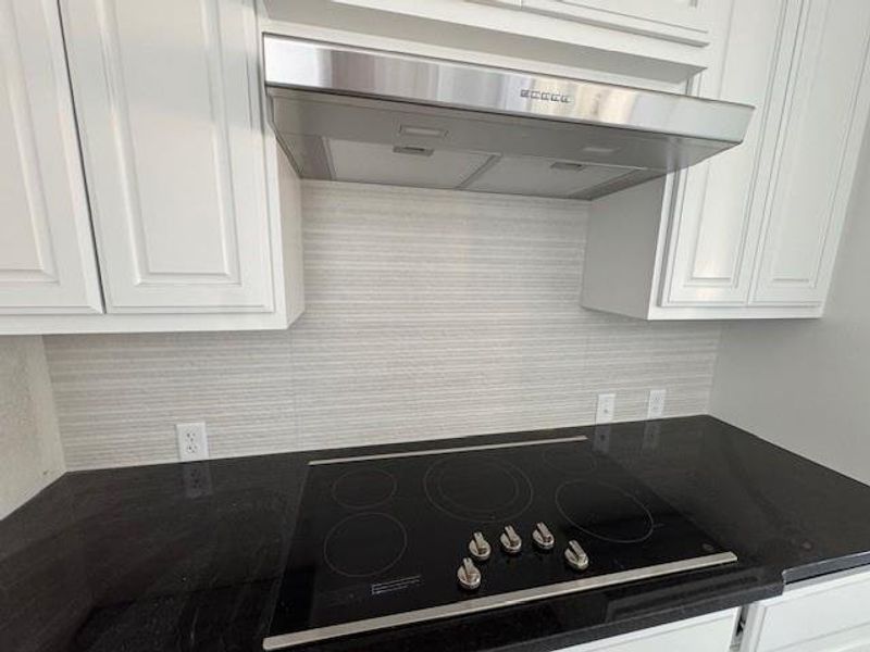 Kitchen view of under cabinet range hood, black electric stovetop, white cabinetry, and dark stone counters