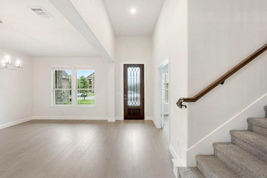 Open entryway with wood front door, light hardwood floors, and carpeted staircase along white walls Open entryway with wood front door, light hardwood floors, and carpeted staircase along white walls