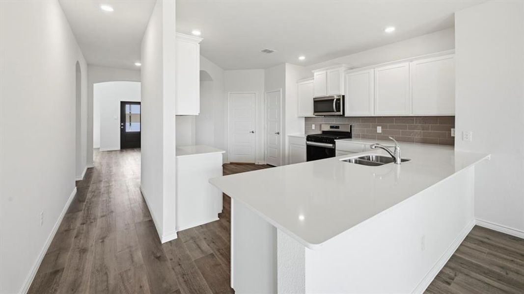 Modern kitchen featuring white cabinetry, a large white countertop island with an undermount sink, a gray subway tile backsplash, and stainless steel appliances including a microwave and gas range Modern kitchen featuring white cabinetry, a large white countertop island with an undermount sink, a gray subway tile backsplash, and stainless steel appliances including a microwave and gas range