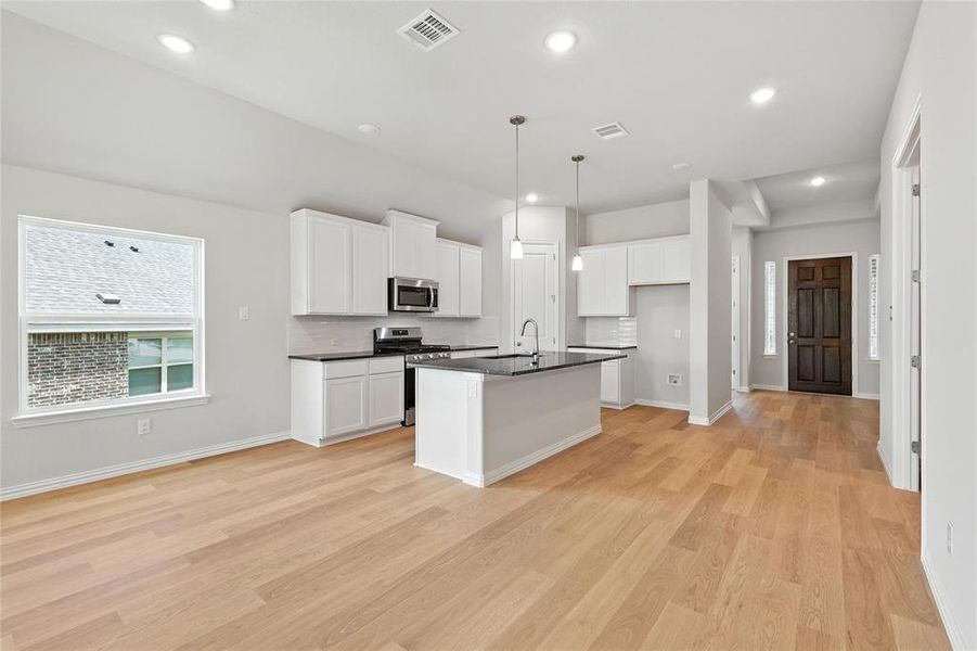Kitchen with appliances with stainless steel finishes, dark countertops, light wood-type flooring, a center island with sink, and backsplash Kitchen with appliances with stainless steel finishes, dark countertops, light wood-type flooring, a center island with sink, and backsplash