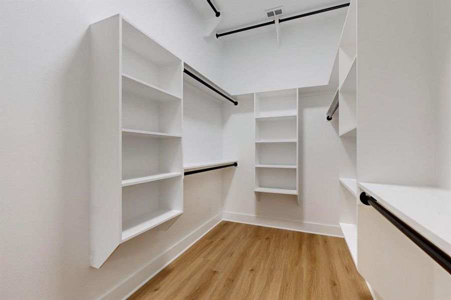 Walk-in closet featuring built-in white shelving, dark-finish hanging rods, and wood-finish flooring Walk-in closet featuring built-in white shelving, dark-finish hanging rods, and wood-finish flooring