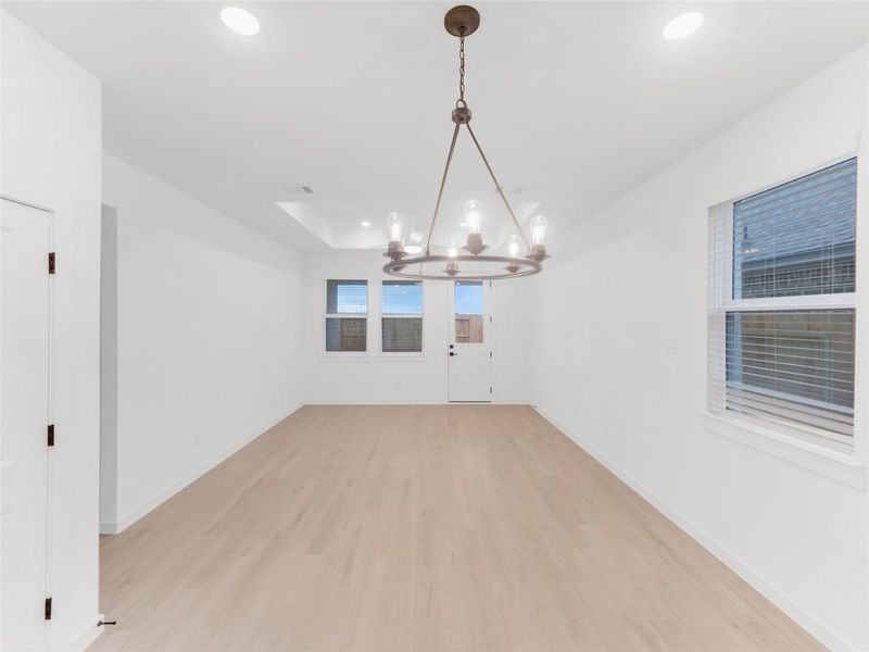 Spacious livingroom with light wood vinyl flooring and white walls. Features a modern chandelier, recessed lighting, large windows, and a door leading outside. Ideal for dining or living area.