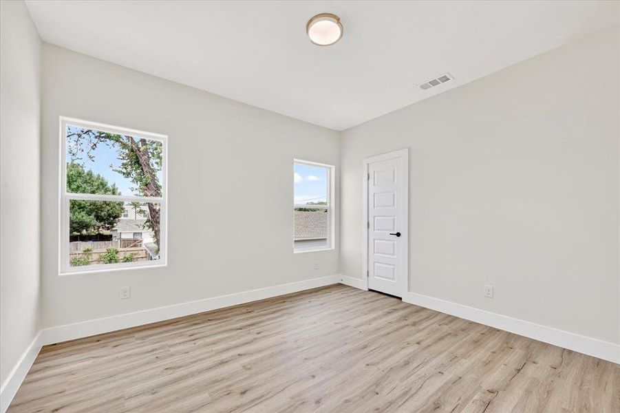 Spacious room featuring wood-finish flooring, light-toned walls, multiple windows providing natural illumination, a contemporary flush-mount ceiling light fixture, and a five-panel interior door with matte black hardware