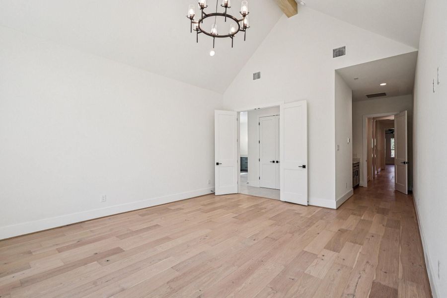 This spacious room features high vaulted ceilings, light wood flooring, and a modern chandelier. It has double doors leading to another area, with additional hallway access. The neutral color scheme enhances the natural light.