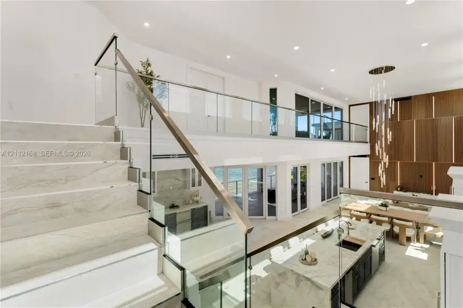 Open-concept interior featuring stone-finish flooring, a glass and metal staircase railing, a multi-level wood-panel wall feature, a stone-topped kitchen island, and multiple glass door systems Open-concept interior featuring stone-finish flooring, a glass and metal staircase railing, a multi-level wood-panel wall feature, a stone-topped kitchen island, and multiple glass door systems