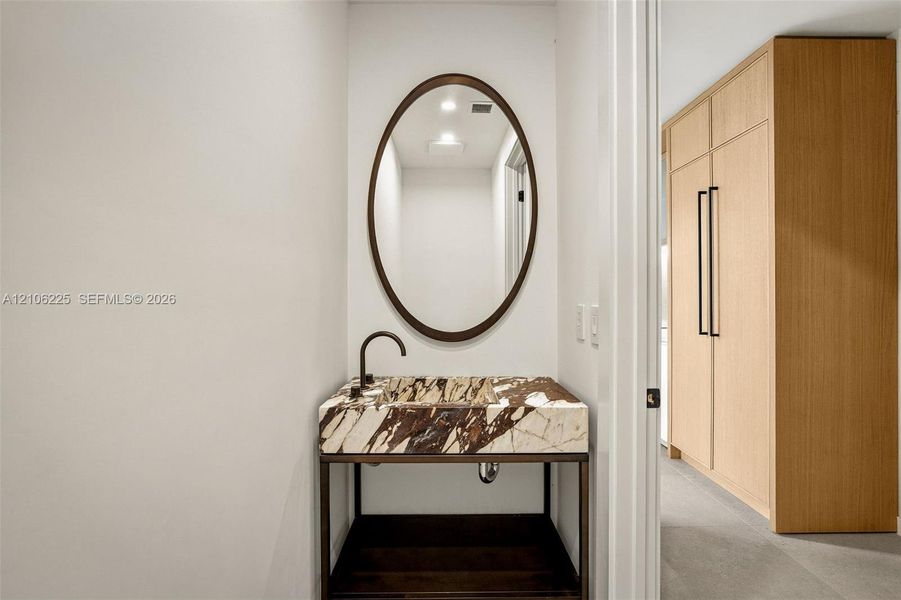 Custom console vanity featuring a stone countertop with integrated basin, oval wall-mounted mirror, matte black faucet, and floor-to-ceiling built-in wood cabinetry Custom console vanity featuring a stone countertop with integrated basin, oval wall-mounted mirror, matte black faucet, and floor-to-ceiling built-in wood cabinetry