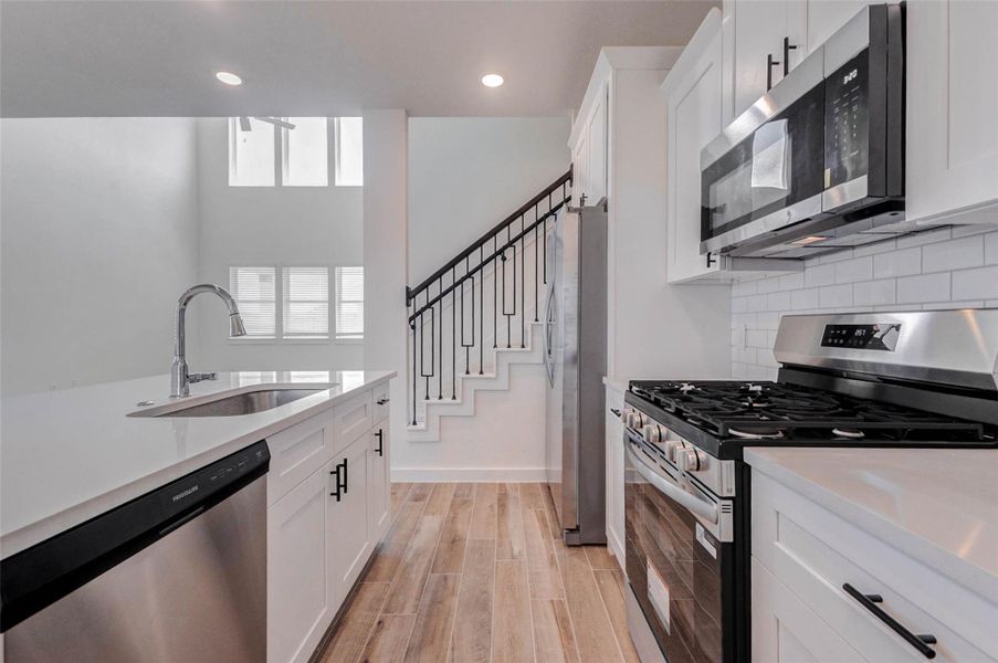 Detailed kitchen view highlighting the large island with sink, quartz-style countertops, stainless dishwasher, gas range, built-in microwave, subway tile backsplash, white shaker-style cabinetry, and black hardware. The kitchen opens toward the living area and staircase, creating a connected layout that is ideal for cooking, hosting, and staying engaged with the household. Detailed kitchen view highlighting the large island with sink, quartz-style countertops, stainless dishwasher, gas range, built-in microwave, subway tile backsplash, white shaker-style cabinetry, and black hardware. The kitchen opens toward the living area and staircase, creating a connected layout that is ideal for cooking, hosting, and staying engaged with the household.