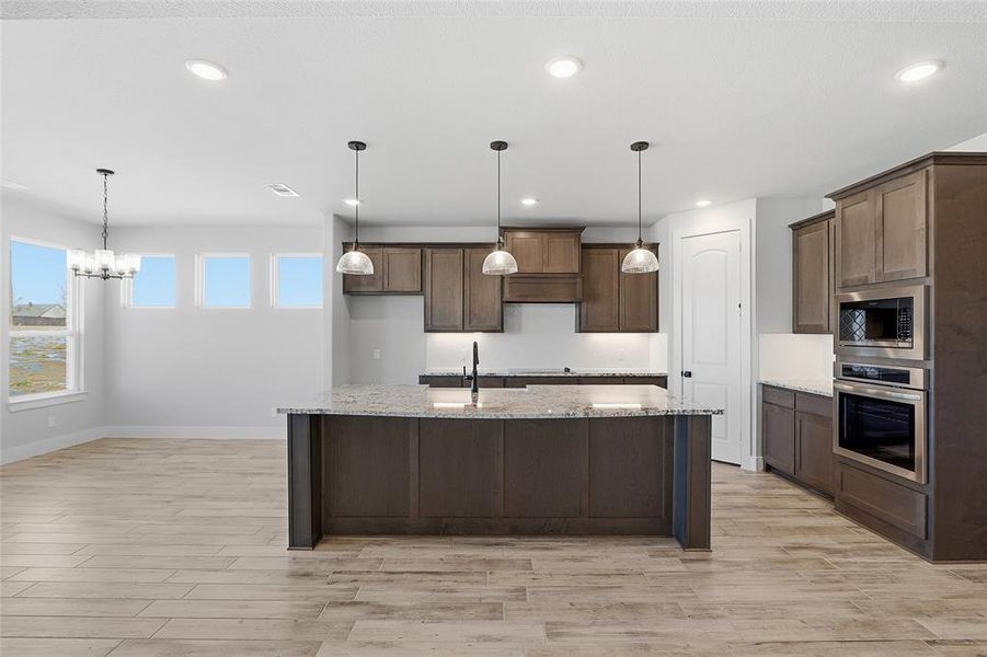 Kitchen with pendant lighting, light stone countertops, stainless steel appliances, light wood-style floors, and an island with sink