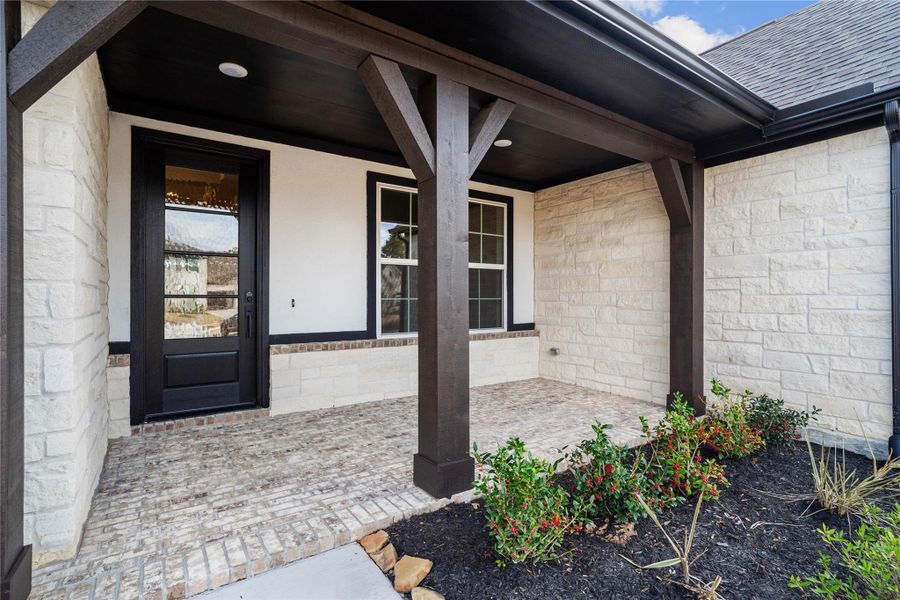 This covered front porch makes a sophisticated first impression, featuring a striking contrast between crisp white chopped stone walls and dark, earth-toned timber beams. The entryway is grounded by a rustic brick-paver floor, leading to a stately black-framed glass front door that perfectly complements the home's modern transitional style.