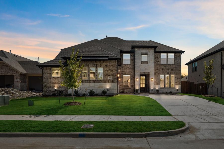 Two-story brick and stone home with dark roof, lit entry, and manicured front lawn at dusk Two-story brick and stone home with dark roof, lit entry, and manicured front lawn at dusk