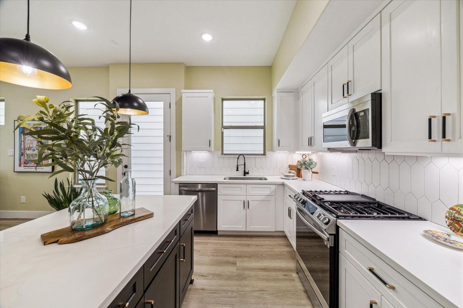 This modern kitchen features sleek white cabinetry, a stylish hexagonal tile backsplash, and stainless steel appliances. The spacious island offers additional prep space and seating, enhanced by elegant pendant lighting. Natural light fills the room, creating a bright and inviting atmosphere.