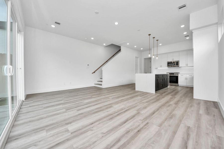 This spacious open-concept living area features modern flooring, large sliding glass doors for natural light, and a sleek kitchen with an island and pendant lighting. A staircase leads to the upper floor, enhancing the contemporary feel.
