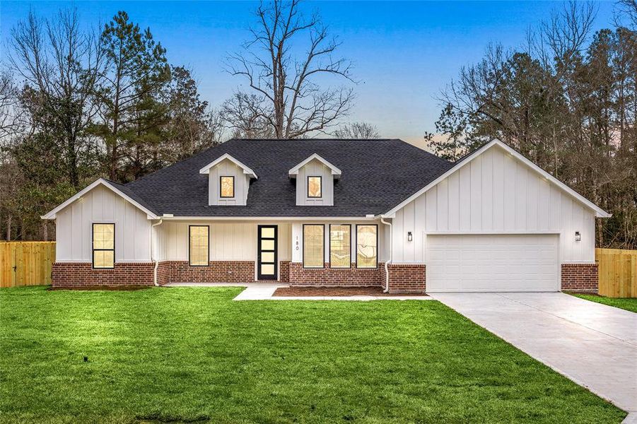 This charming home features a modern farmhouse design with a white exterior, brick accents, and a black roof. It includes a welcoming front porch, a two-car garage, and a spacious driveway. The property is surrounded by lush greenery and mature trees, offering a serene setting.