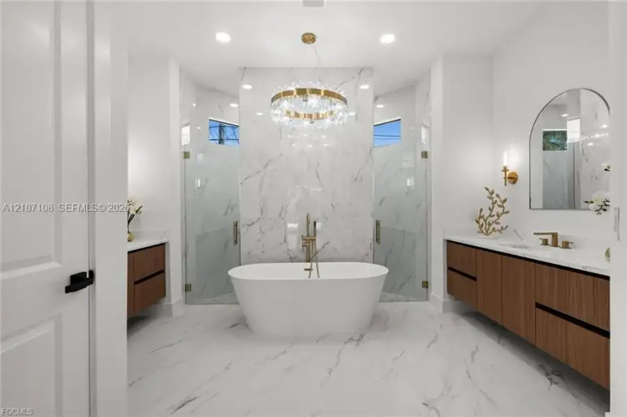 Freestanding white soaking tub, dual floating wood-finish vanities, wall-to-wall stone-surround shower, gold-tone plumbing fixtures, and a circular crystal chandelier