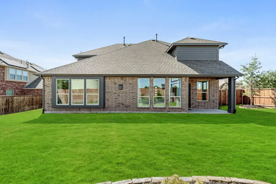 Rear exterior of brick home with covered patio, large windows, and fenced backyard with green lawn