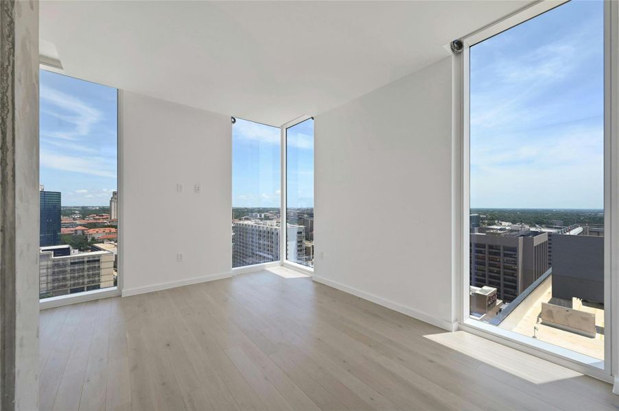 Stunning primary bedroom featuring dramatic floor-to-ceiling corner windows that flood the space with natural light and showcase panoramic downtown views. Light-toned hardwood floors and clean modern finishes enhance the bright, minimalist aesthetic—perfect for a serene city retreat.