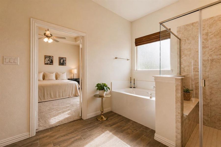 Bright bathroom featuring a built-in soaking tub with a window, a glass-enclosed shower with tile surround, and wood-finish flooring