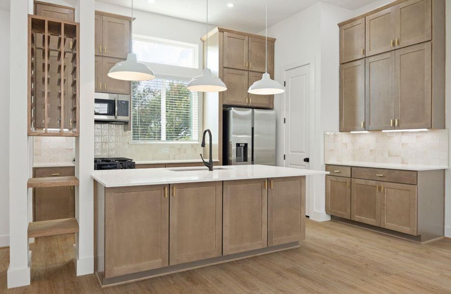 Modern kitchen with a generous island, sleek quartz counters, warm cabinetry, brass hardware, under-cabinet lighting, stainless steel appliances, and a soft neutral tile backsplash. The built-in wine/display storage adds a custom touch, while pendant lights create a polished focal point above the island.