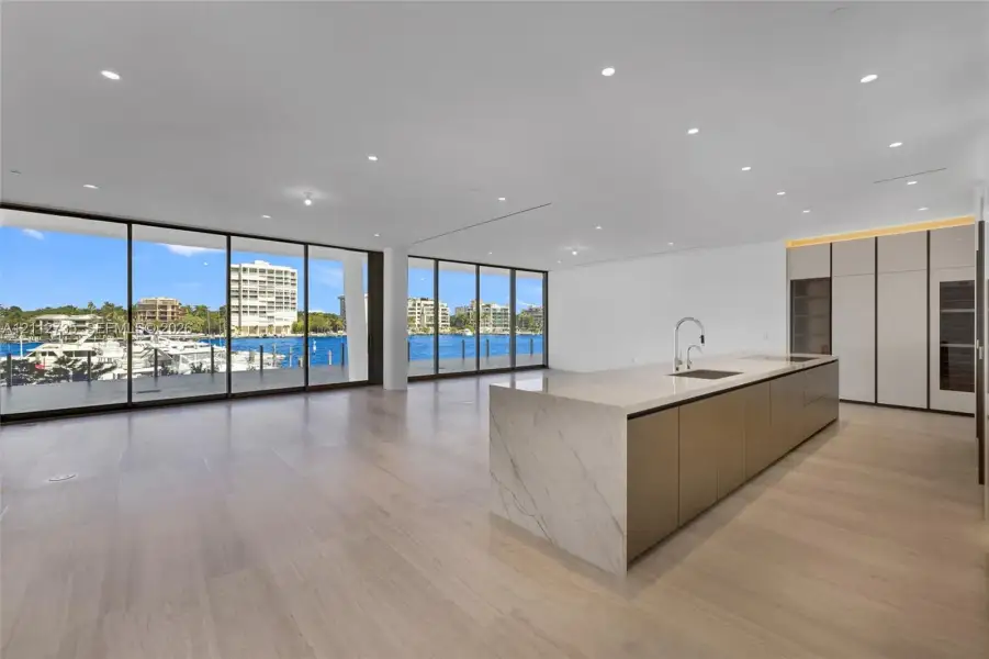 Open-concept interior featuring floor-to-ceiling glass walls, expansive kitchen island with a stone-finish waterfall countertop, built-in floor-to-ceiling cabinetry, and light-toned wood-finish flooring