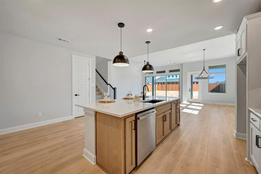 Kitchen with light wood-style flooring, stainless steel dishwasher, a kitchen island with sink, hanging light fixtures, and light stone counters