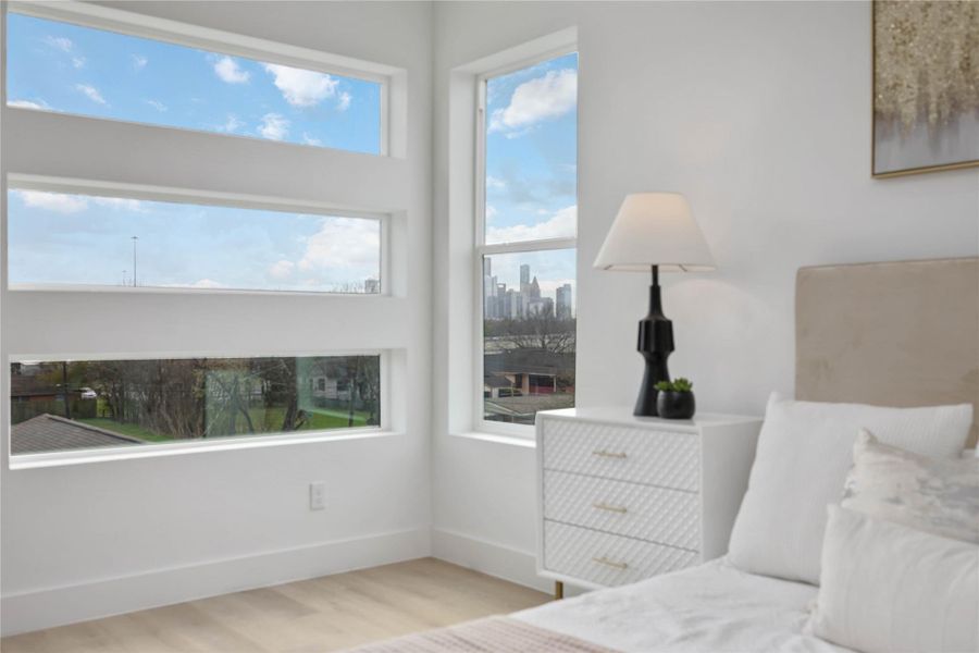 Large modern picture windows frame beautiful natural views and fill the bedroom with abundant daylight, highlighting the clean white walls, warm flooring, and contemporary designer touches throughout the space.