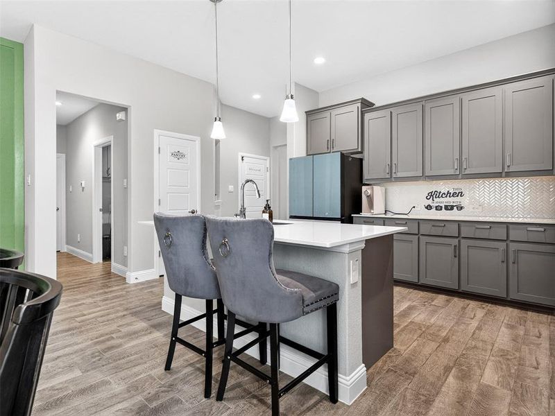 Central kitchen island with white countertop, pendant lighting, contemporary gray cabinetry, herringbone tile backsplash, and wood-finish flooring Central kitchen island with white countertop, pendant lighting, contemporary gray cabinetry, herringbone tile backsplash, and wood-finish flooring