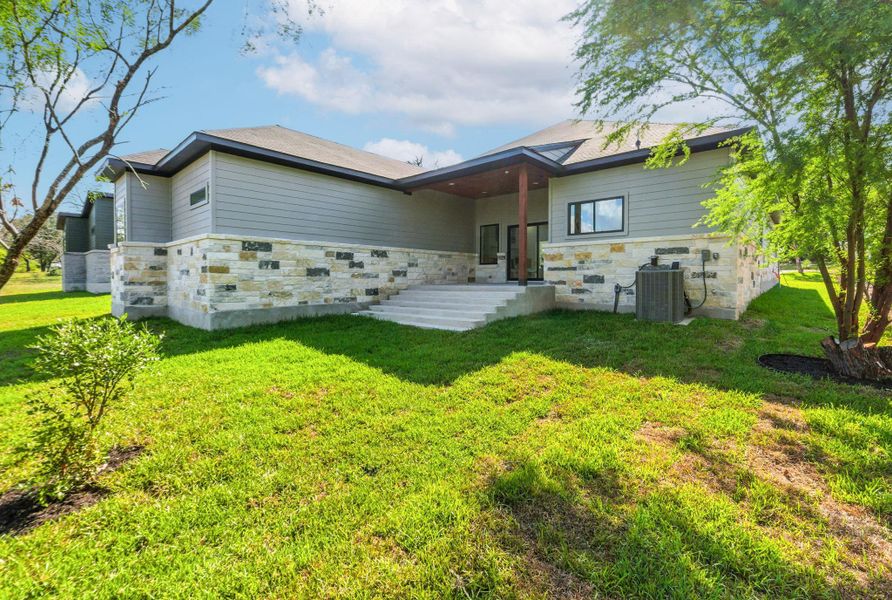 A spacious backyard with a covered patio featuring a ceiling fan, stone accents, and wide steps leading to a lush green lawn, surrounded by mature trees and natural landscaping