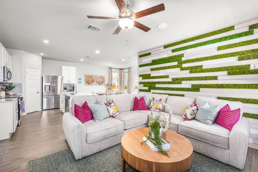 Image of living room and kitchen with wood flooring, a greenery and wood paneling accent wall, and a tan couch with pink a blue pillows.
