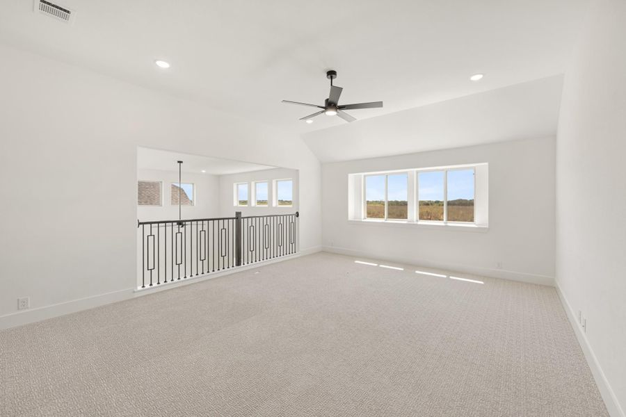 Open upstairs bonus room with vaulted ceiling, ceiling fan, carpet flooring, and black iron stair railing overlooking lower level.