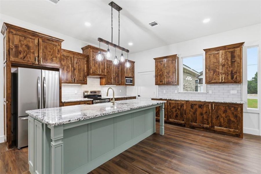 Spacious kitchen featuring a large island with granite countertop, wood-finish flooring, undermount sink with a gooseneck faucet, white subway tile backsplash, and dark wood cabinetry Spacious kitchen featuring a large island with granite countertop, wood-finish flooring, undermount sink with a gooseneck faucet, white subway tile backsplash, and dark wood cabinetry